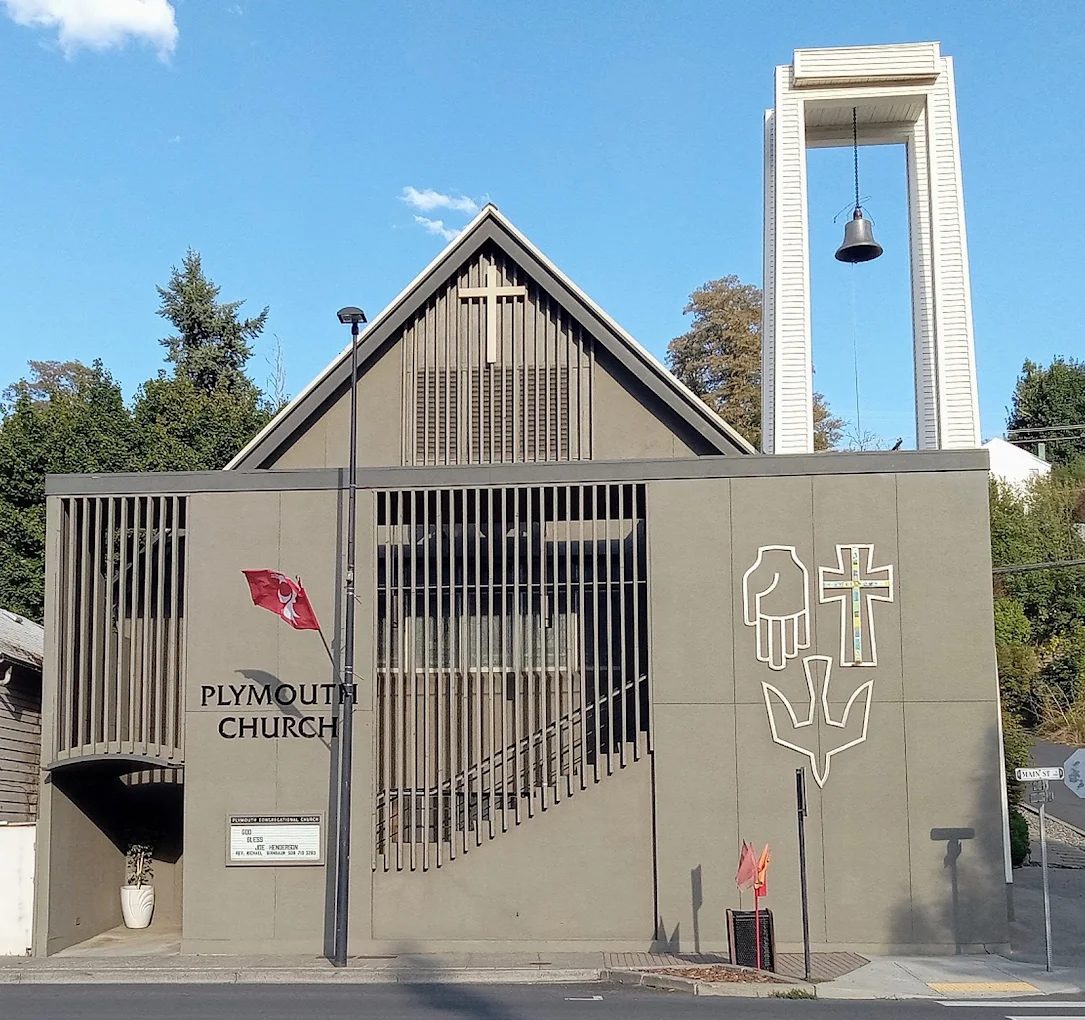 Plymouth Church, a mid-century modern building at 321 S Main St in Colfax, Washington, with a tall freestanding white bell tower and carved symbols of a hand, cross, and dove on its taupe facade.